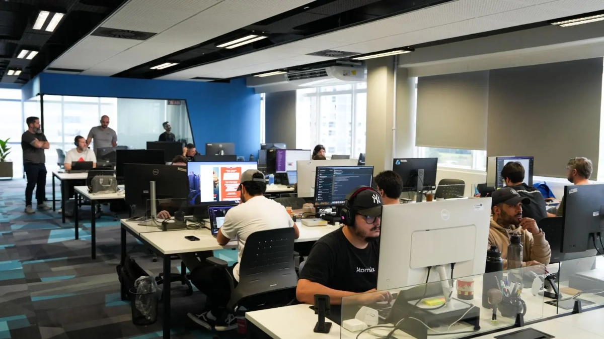 Vista ampla de um escritório moderno e movimentado com várias pessoas sentadas em mesas trabalhando em computadores e outras pessoas em pé conversando ao fundo. O escritório tem carpetes coloridos e grandes janelas.