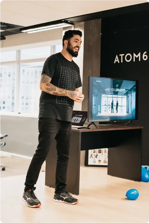 Um homem barbudo com tatuagens nos braços, vestindo uma camiseta preta, sorri enquanto está de pé ao lado de uma televisão que exibe uma apresentação com o texto 'ARE YOU READY?'. Um balão azul está no chão à direita.