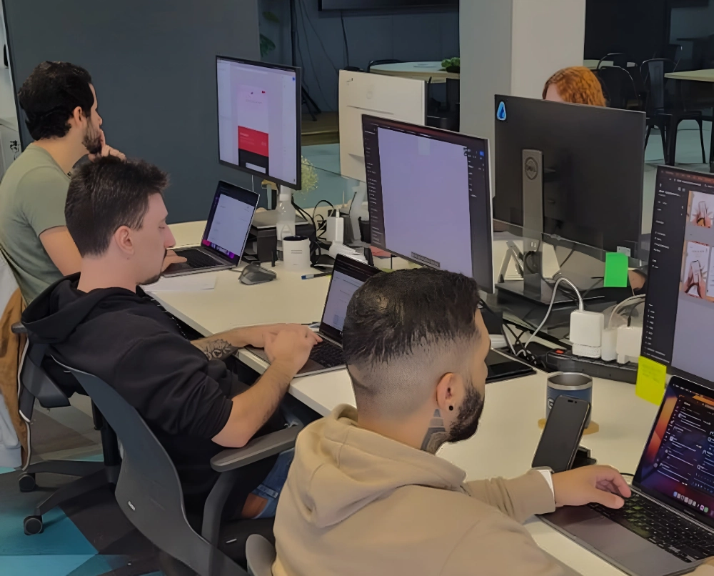 Quatro pessoas em um escritório, sentadas em mesas com computadores. Um homem de barba e capuz está de costas, outro de perfil usando óculos, e uma pessoa com cabelo ruivo sentada em frente a um monitor.