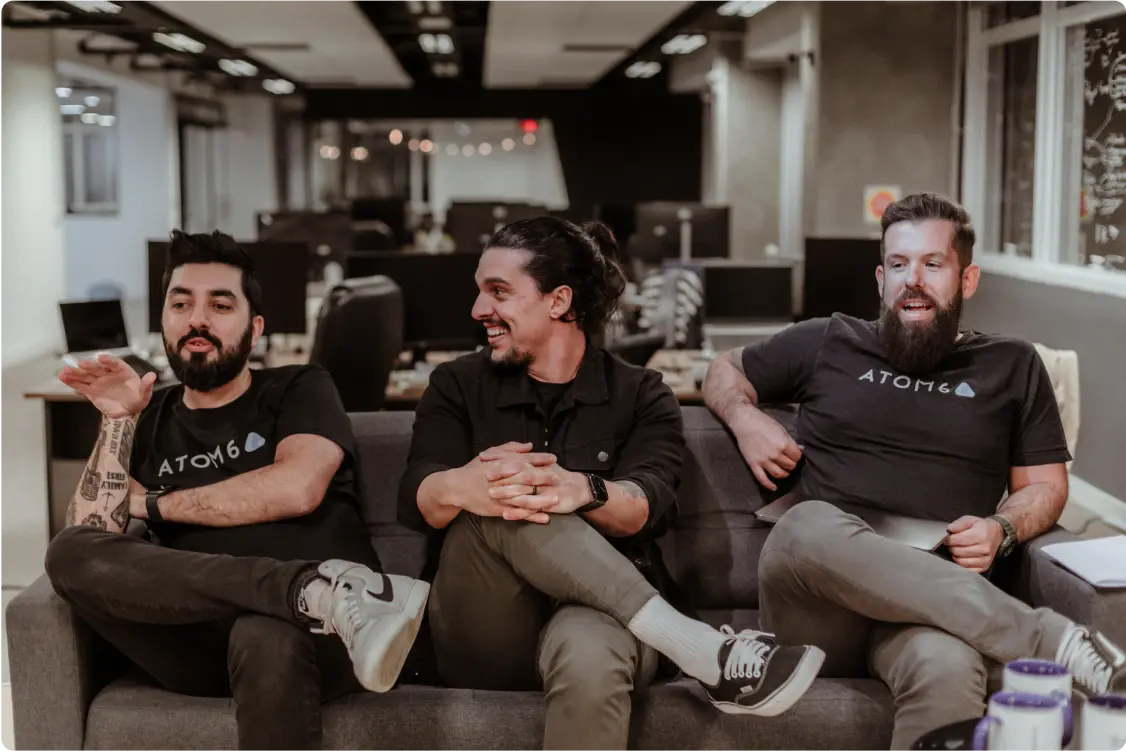 Três homens com barbas e camisetas pretas sentados confortavelmente em um sofá em um escritório, conversando e rindo. Um deles gesticula com a mão.